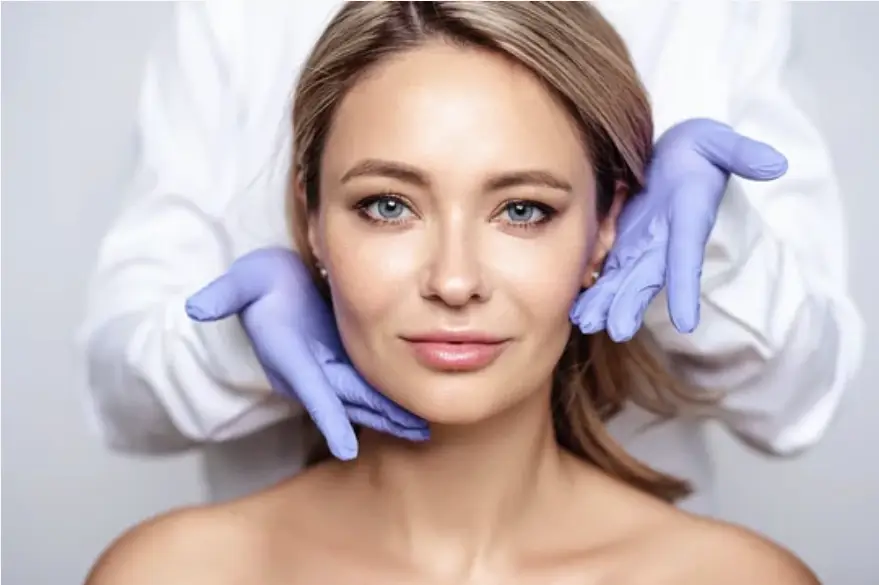 A doctor wearing blue gloves gently touches a woman's face, holding her jawline. The woman has light hair and blue eyes, and she is looking directly at the camera. The doctor is in the background, out of focus, wearing a white coat.